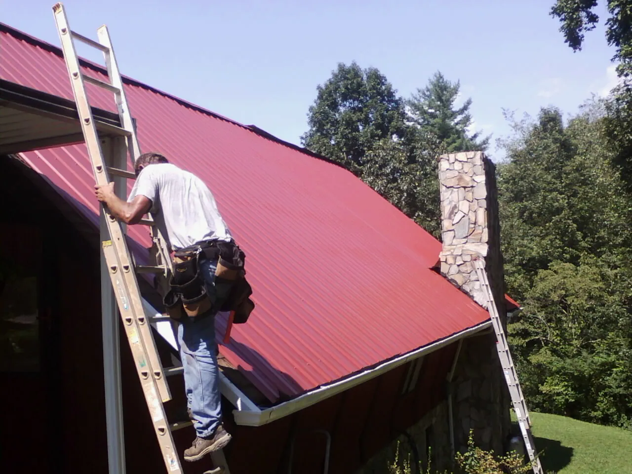 Roofer on a ladder beside a red metal roof and stone chimney — Soffit & Fascia Repair by Roofing Brothers in West Cornwall
